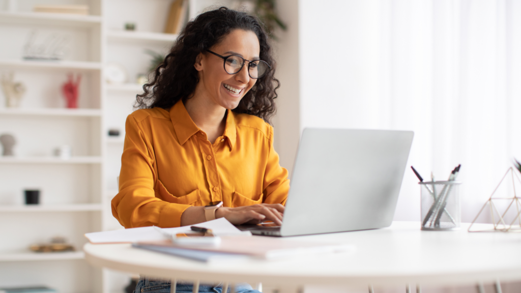 A woman wearing glasses sits at a table, focused on her laptop while working or studying.