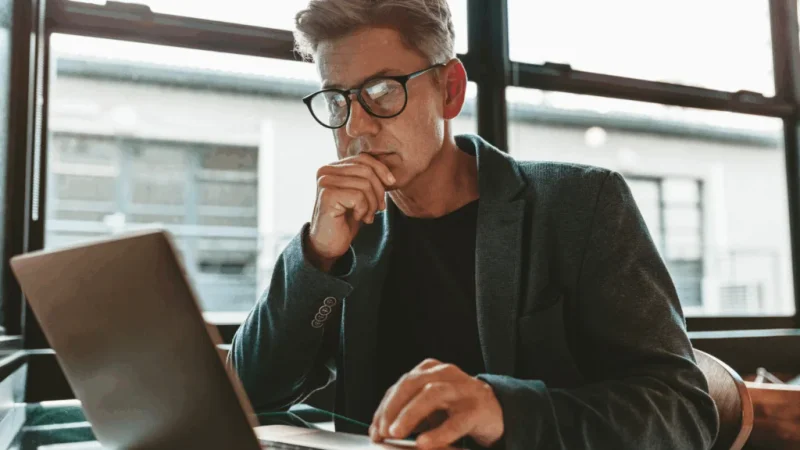 A man in glasses is looking at a laptop screen.
