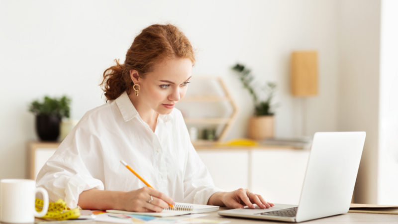 A woman seated at a table, working on a laptop with a pen in hand, focused on her task.