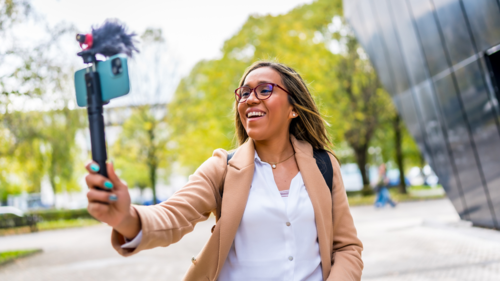 A smiling woman walking outside and vlogging using a mobile phone she’s holding on a tripod in her hand.