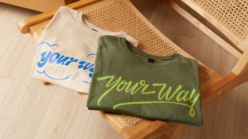 Two folded “Your way” t-shirts on a wooden chair. One t-shirt is cream with blue text, the second t-shirt is khaki with green text.
