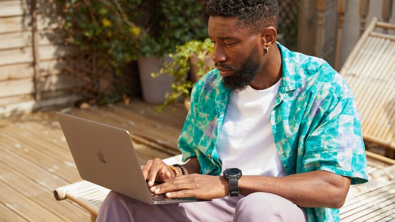 A man working on a laptop outdoors, searching how to digitize photos, sitting on a lounge chair, wearing a colorful blue shirt.