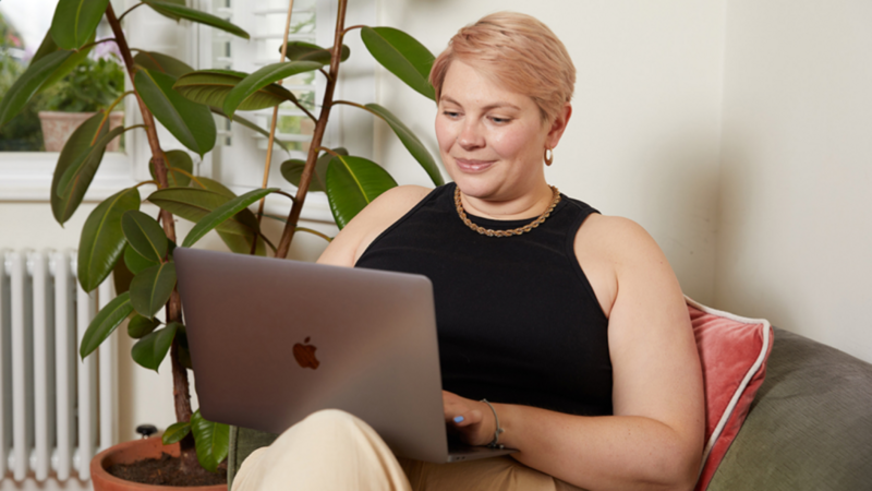 A woman with short hair smiles while working on a laptop. She sits on a couch beside a potted plant.