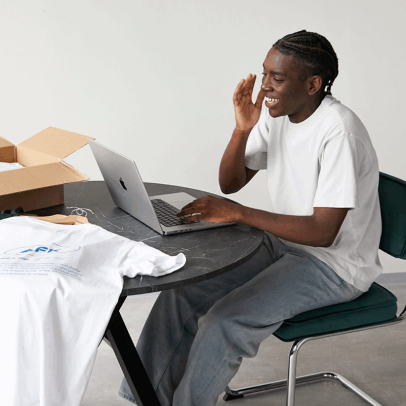 A smiling young man is sitting at a table using a laptop. Custom t-shirts are placed near the laptop.