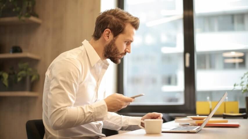 A man sitting at a desk and looking at a laptop screen while holding a mobile phone.