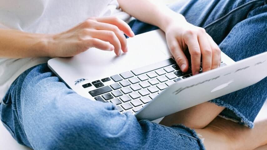 A man in blue jeans and a white t-shirt holding a laptop on his lap and typing.