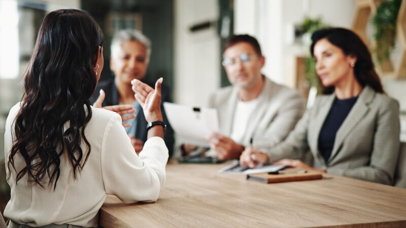 Woman gesturing while speaking to three colleagues seated at a table during a meeting.