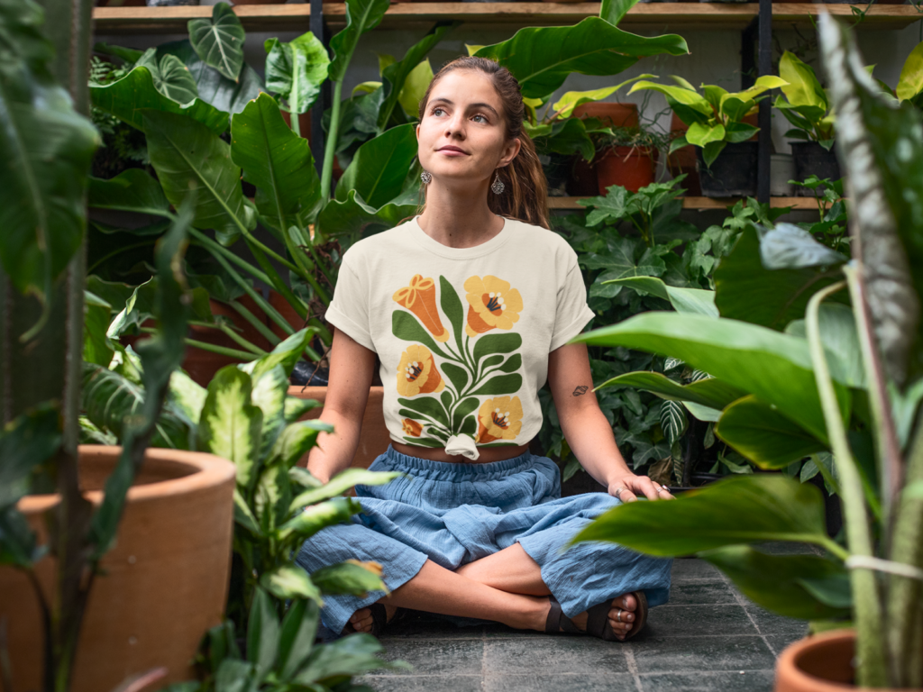 A woman sits cross-legged in a lush greenhouse, surrounded by vibrant plants. She wears a floral t-shirt and blue skirt.