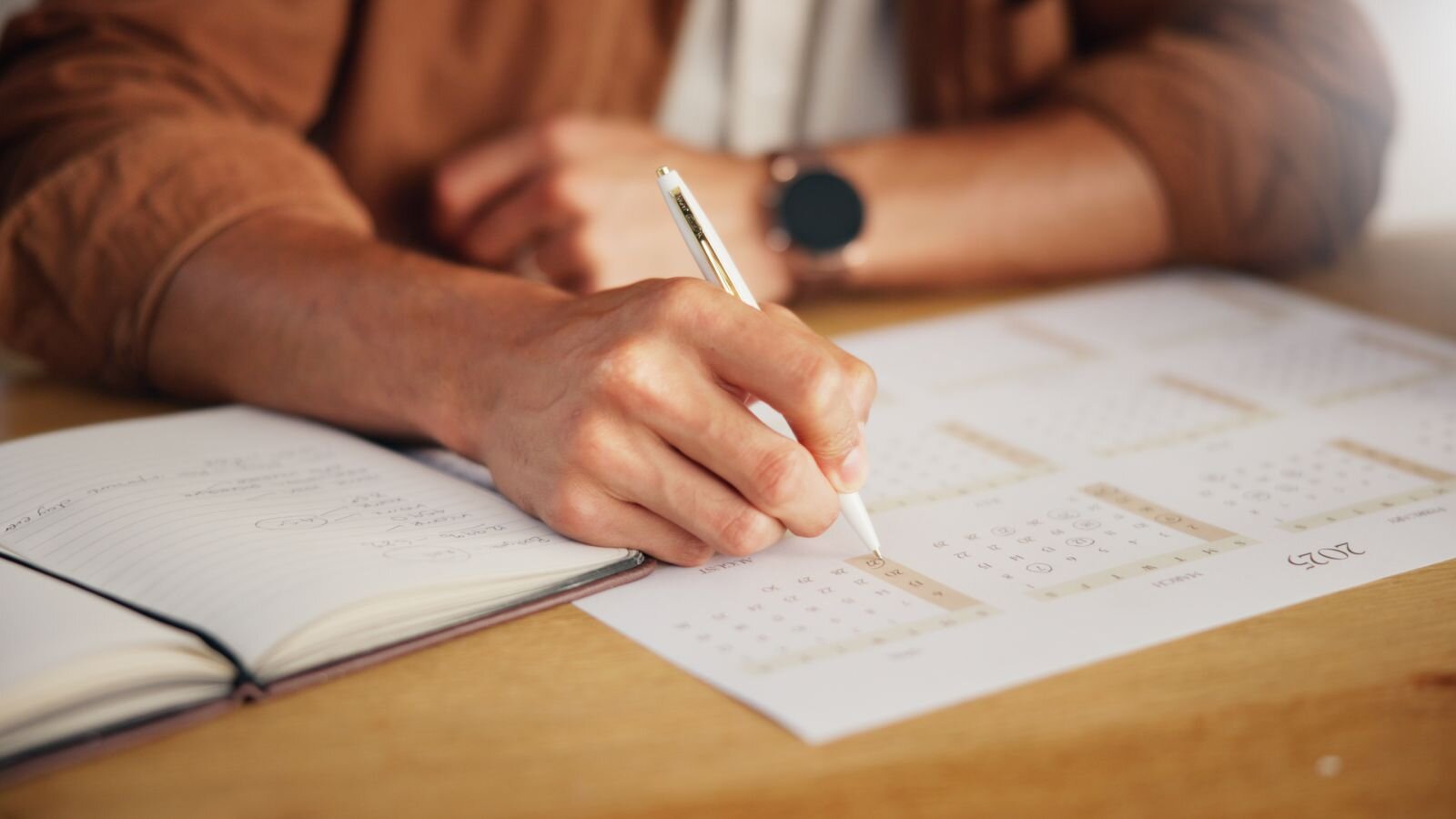 A person writing on a calendar while sitting at a wooden table, with an open notebook nearby.
