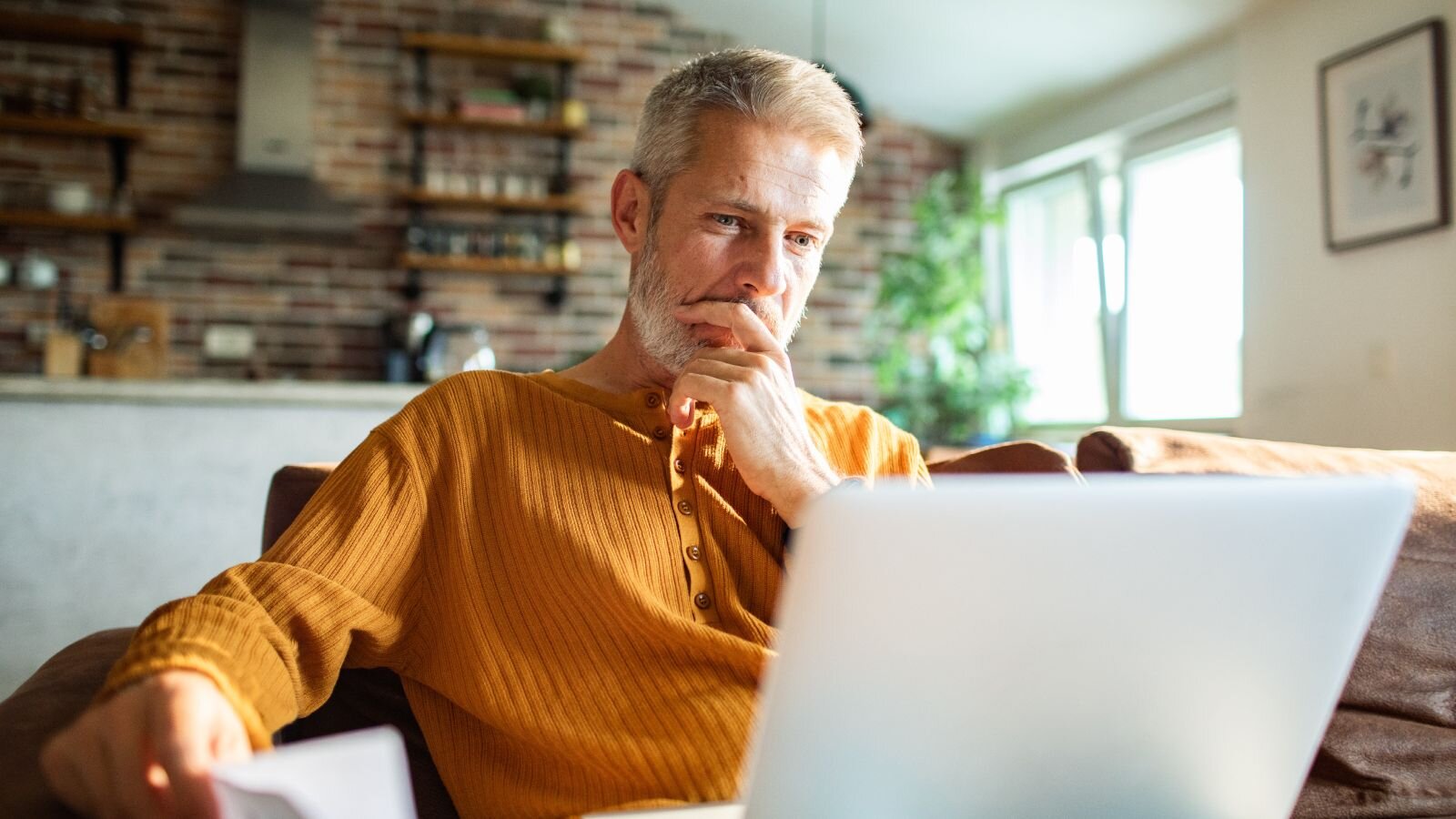 A man sitting on a couch, looking thoughtfully at a laptop, with sunlight coming through the windows in a cozy room.