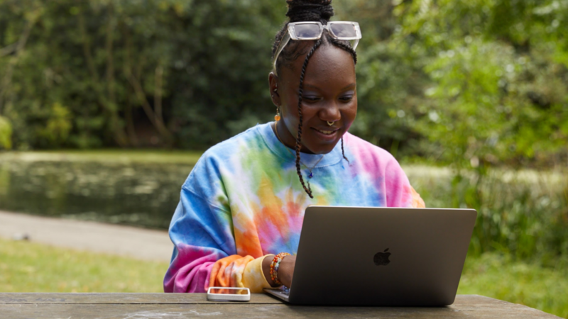 A young woman sitting at a table outdoors, wearing a colorful tie-dye sweatshirt, using a laptop, and smiling.