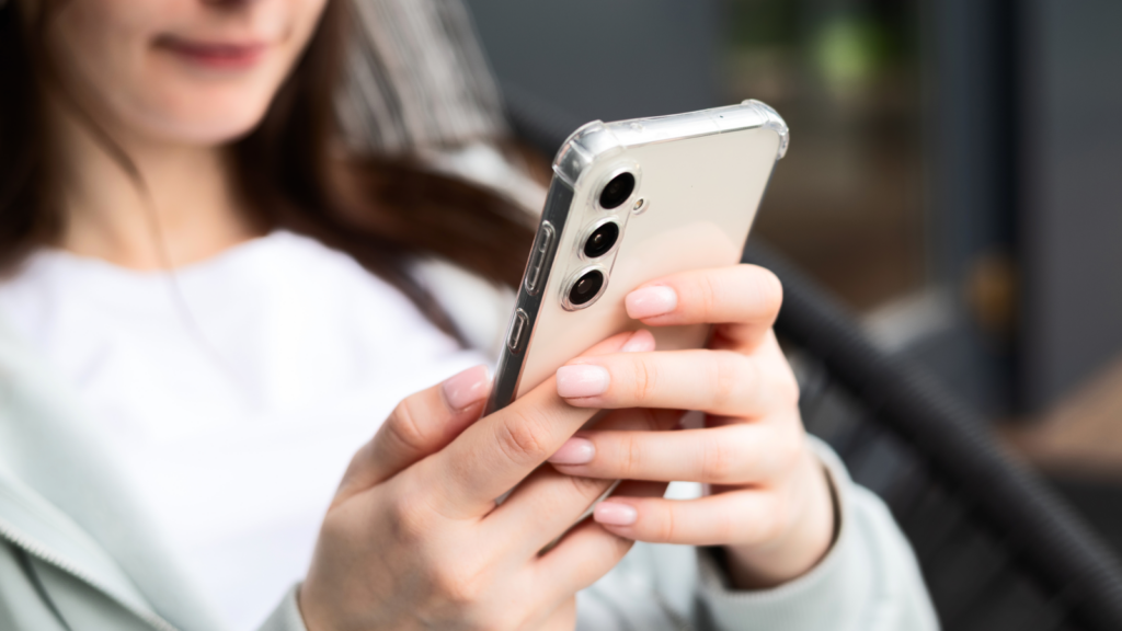 A woman in a casual outfit holds a smartphone with three rear cameras, focusing on the screen.