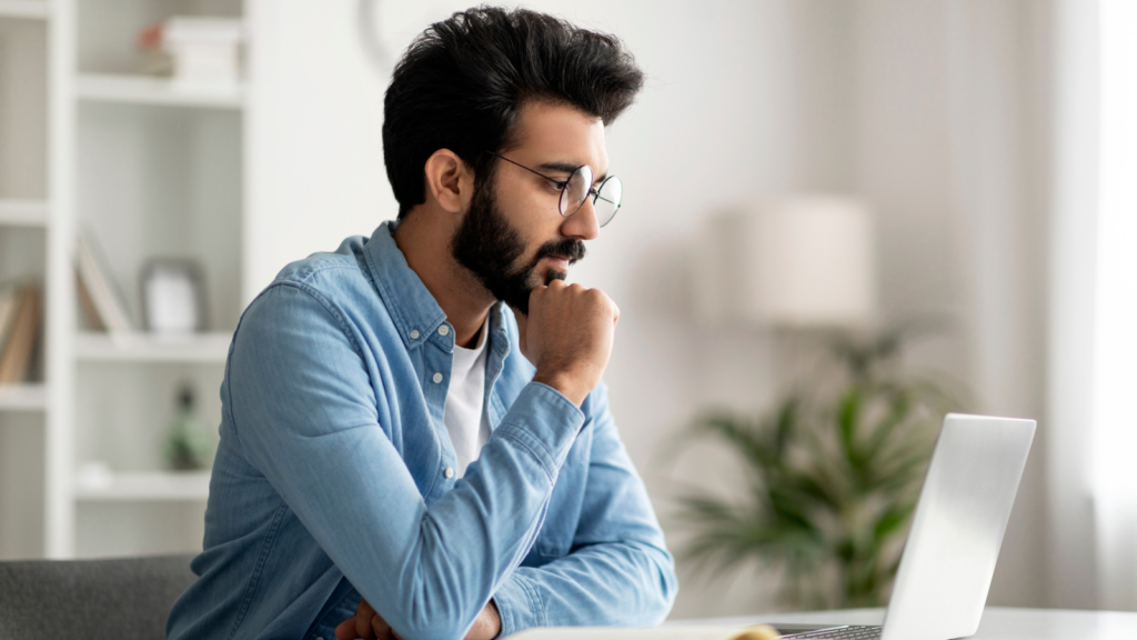 A man in glasses and a jeans shirt, sitting at a desk and looking at a laptop screen.