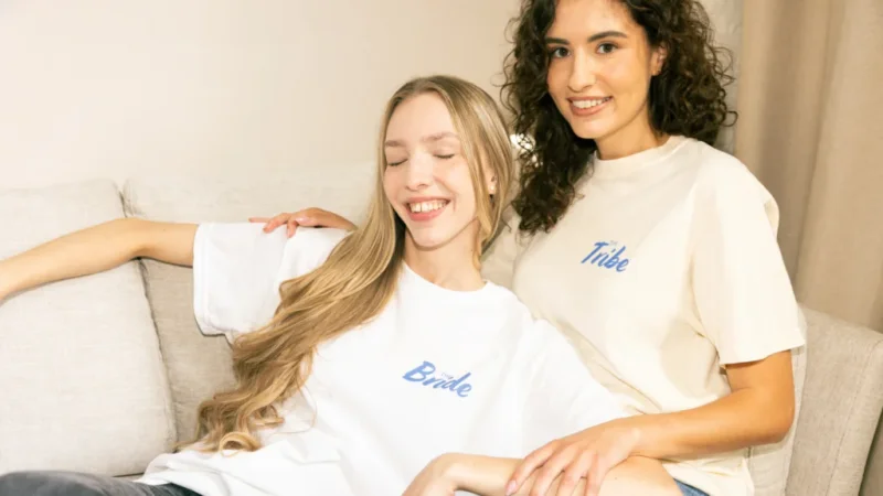 Two women are sitting on a couch, both smiling. One wears a "Bride" t-shirt, the other a "Tribe" t-shirt.