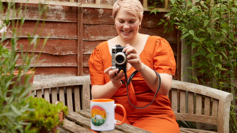 A woman with short blonde hair, wearing an orange dress, is taking a picture of a colorful mug using a digital camera. Home garden in the background.