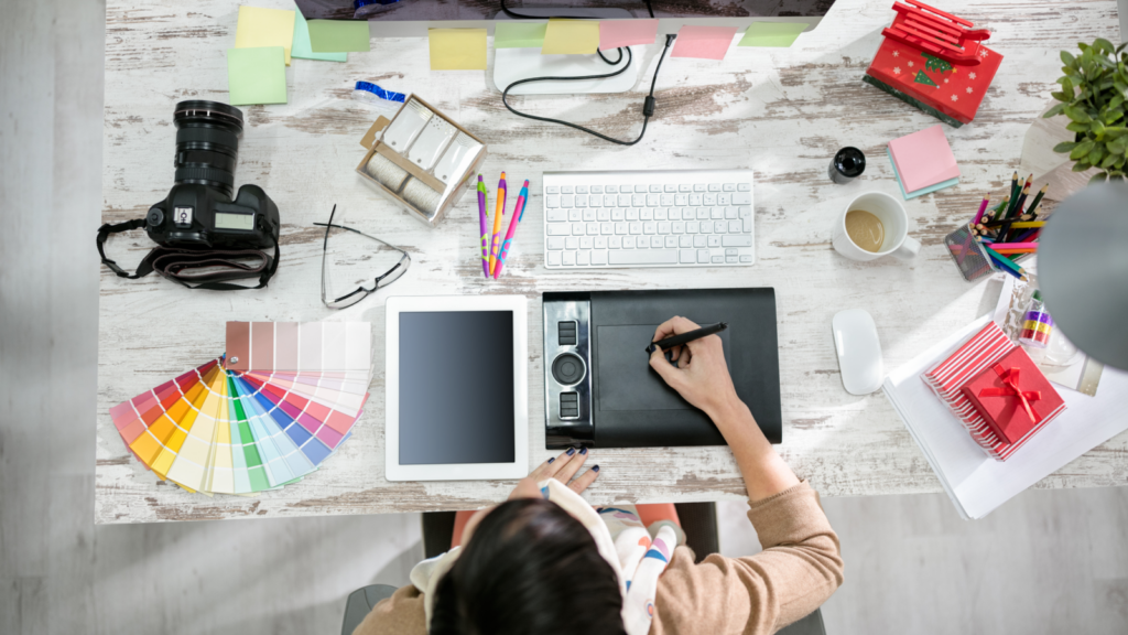 A woman focused on her laptop, surrounded by a tablet and various work items, engaged in graphic design tasks.