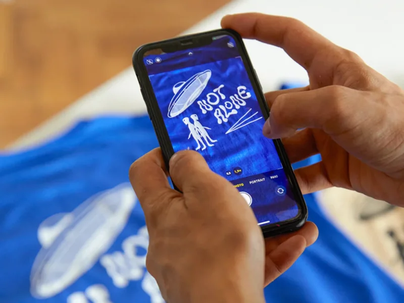 A person holds a smartphone capturing a photo of a blue shirt with a UFO and aliens, featuring the text "Not Alone”.