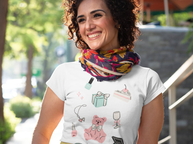 Smiling woman outdoors, wearing a white t-shirt with colorful doodles of makeup, a gift box, cake, and a scarf.