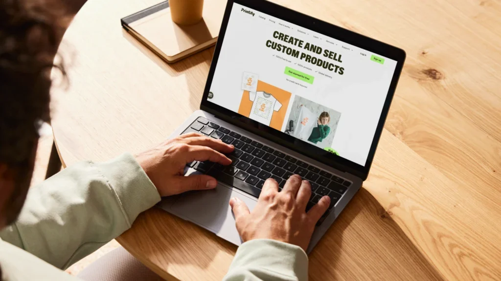 A person is sitting at a table, using a laptop with Printify’s homepage on the screen as an example of how to make money from your phone.