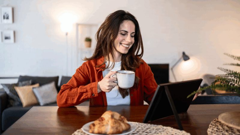 A smiling woman is sitting at a table, drinking coffee and using a tablet to find out when Black Friday is.