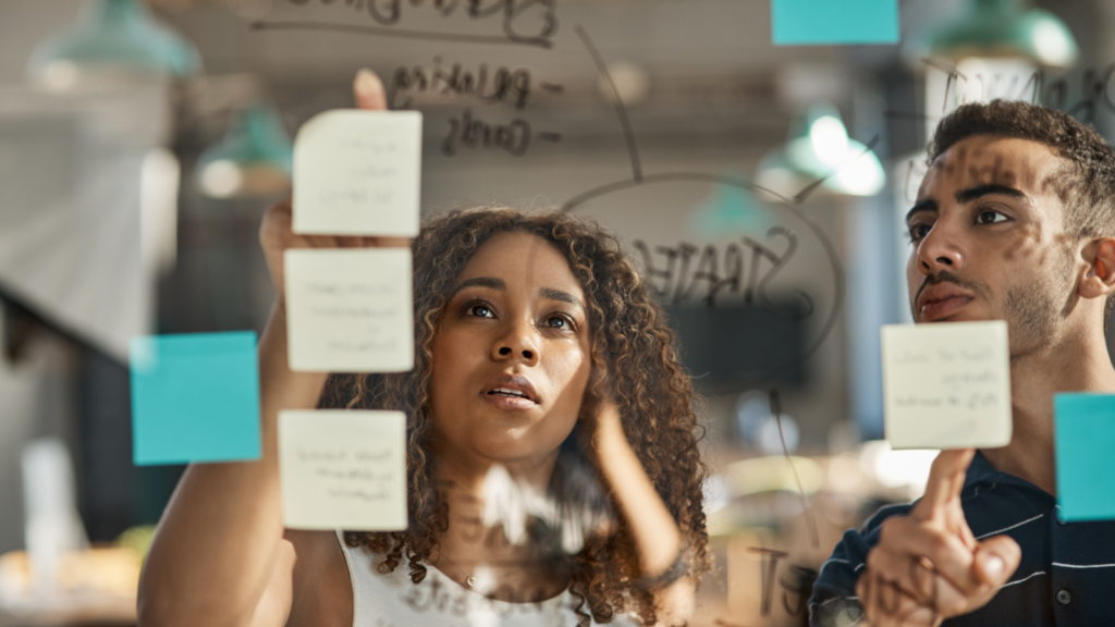 Two individuals examining colorful sticky notes arranged on a transparent glass wall in a collaborative workspace.