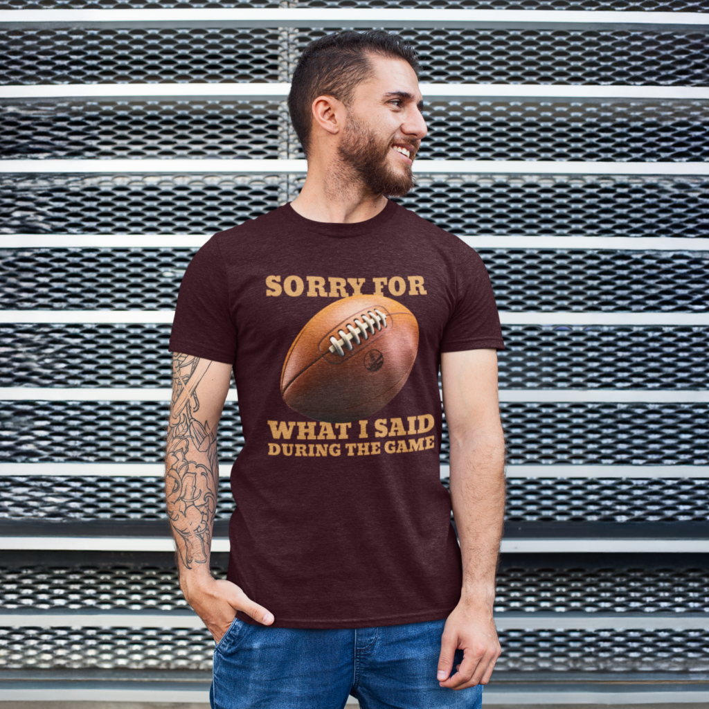 A man with a beard, smiling, wearing a maroon t-shirt with a football graphic and text, "Sorry for what I said during the game".