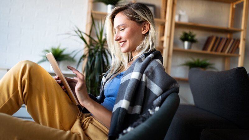 A woman sitting comfortably in a chair, wearing a blue shirt and yellow pants, using a tablet while wrapped in a blanket.