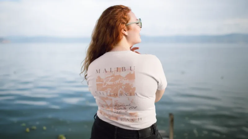 A backshot of a woman with red hair and in glasses wearing a white “Malibu, California” t-shirt. Sea in the background.
