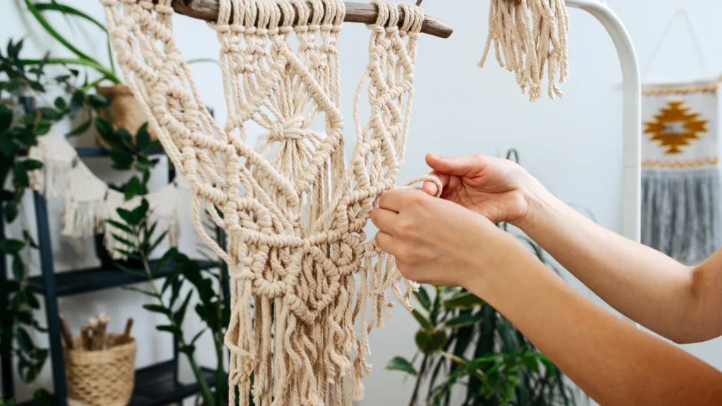 Close-up of hands crafting a macram�� wall hanging with intricate patterns. The setting is a bright room with plants.