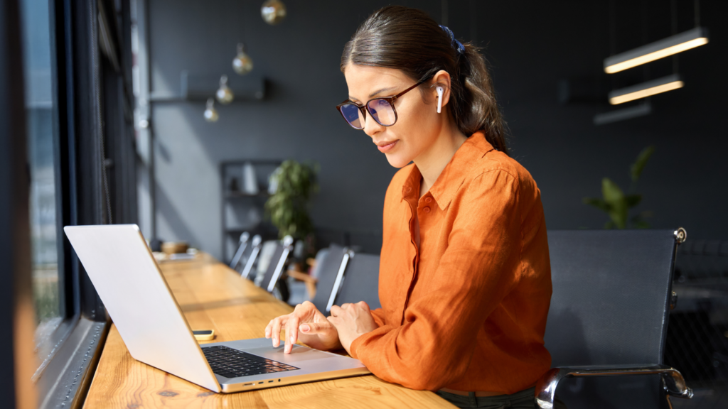A woman in an orange blouse with glasses and earbuds works on a laptop at a wooden table in a modern cafe.