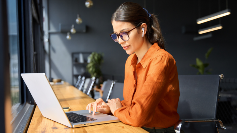 A woman in an orange blouse with glasses and earbuds works on a laptop at a wooden table in a modern cafe.
