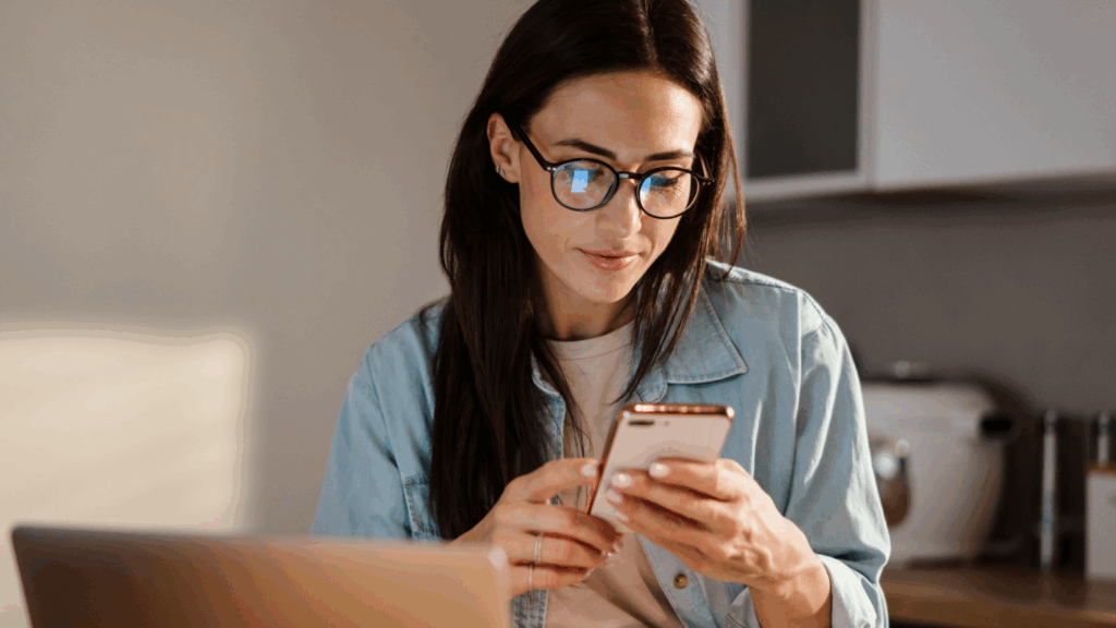 A young woman in glasses is using a mobile phone while sitting in front of a laptop.