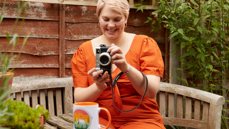 A woman in an orange dress is taking a picture of a colorful mug in an outdoor garden area.