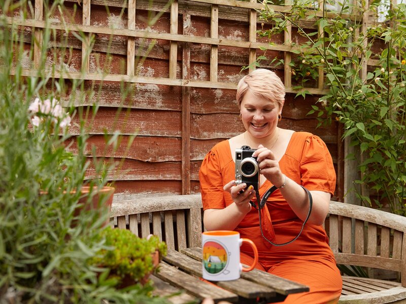 A woman with short blonde hair, wearing an orange dress, is taking a picture of a colorful mug using a camera. Home garden background.