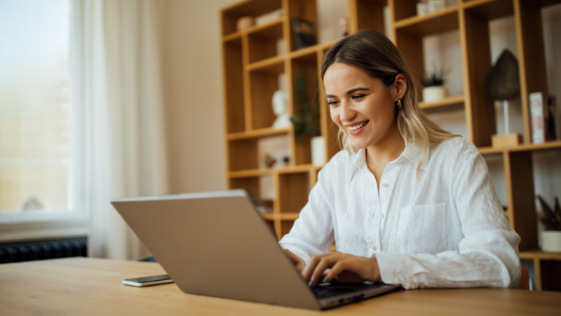 A smiling woman in a white shirt sitting in a home office, using a laptop to look for Wix alternatives.