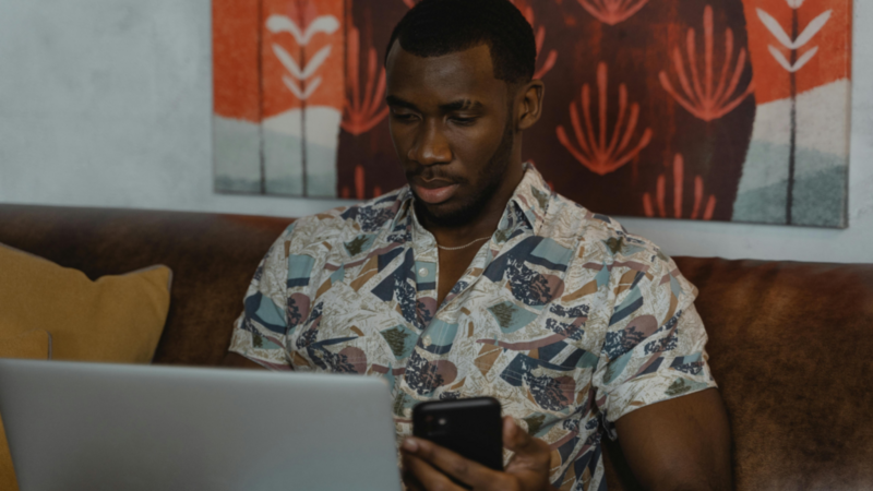 A man sitting on a couch in front of a laptop and holding a mobile phone in his hand.