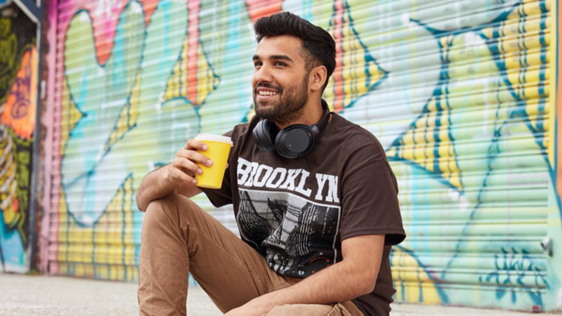 A smiling man is sitting outside with a takeaway coffee cup, wearing a brown “Brooklyn” t-shirt. Graffiti in the background.
