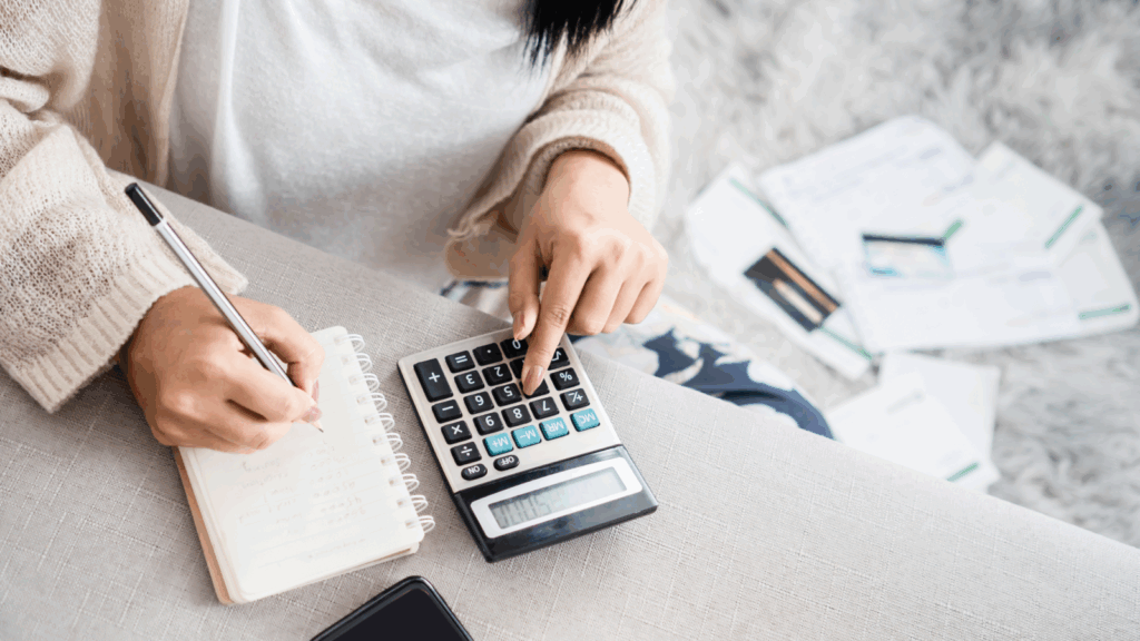 A woman uses a calculator and takes notes in a notebook. Bills and documents scatter on a soft rug.