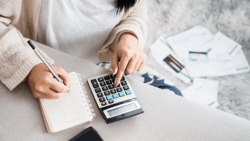 A woman uses a calculator and takes notes in a notebook. Bills and documents scatter on a soft rug.
