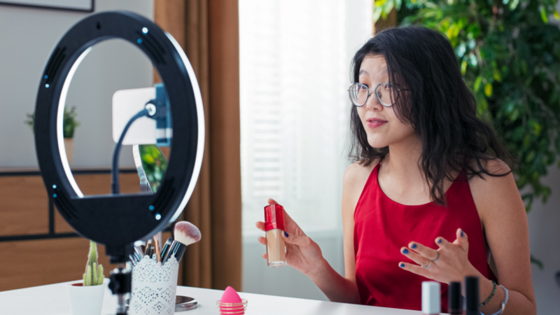 A woman in glasses and a red blouse is presenting beauty products during a livestream, using a smartphone and ring light to stream.