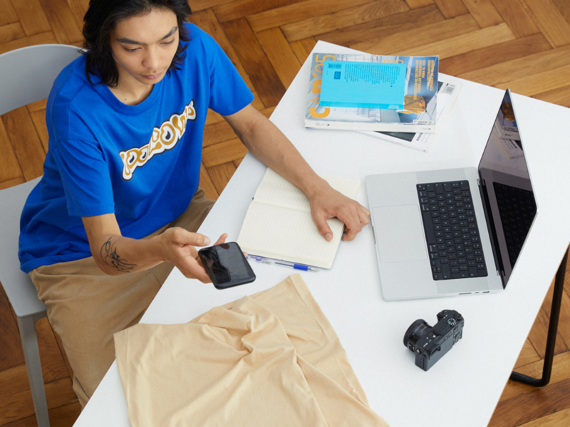 A young man taking a picture of a beige t-shirts with his smartphone. Laptop, camera and notebook are placed around the table.