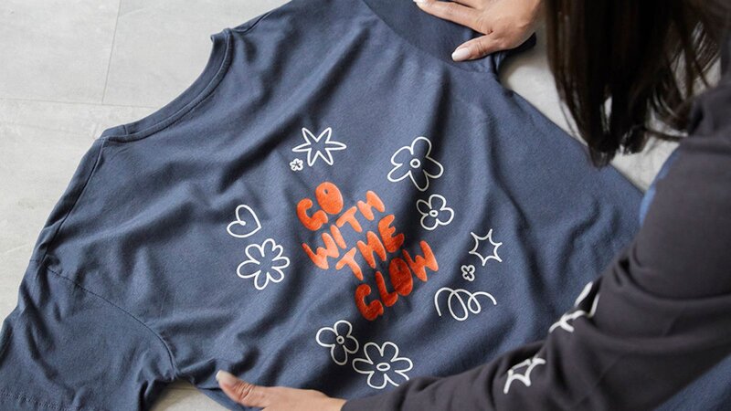 A person stretching out a blue “Go with the glow” t-shirt on a surface.