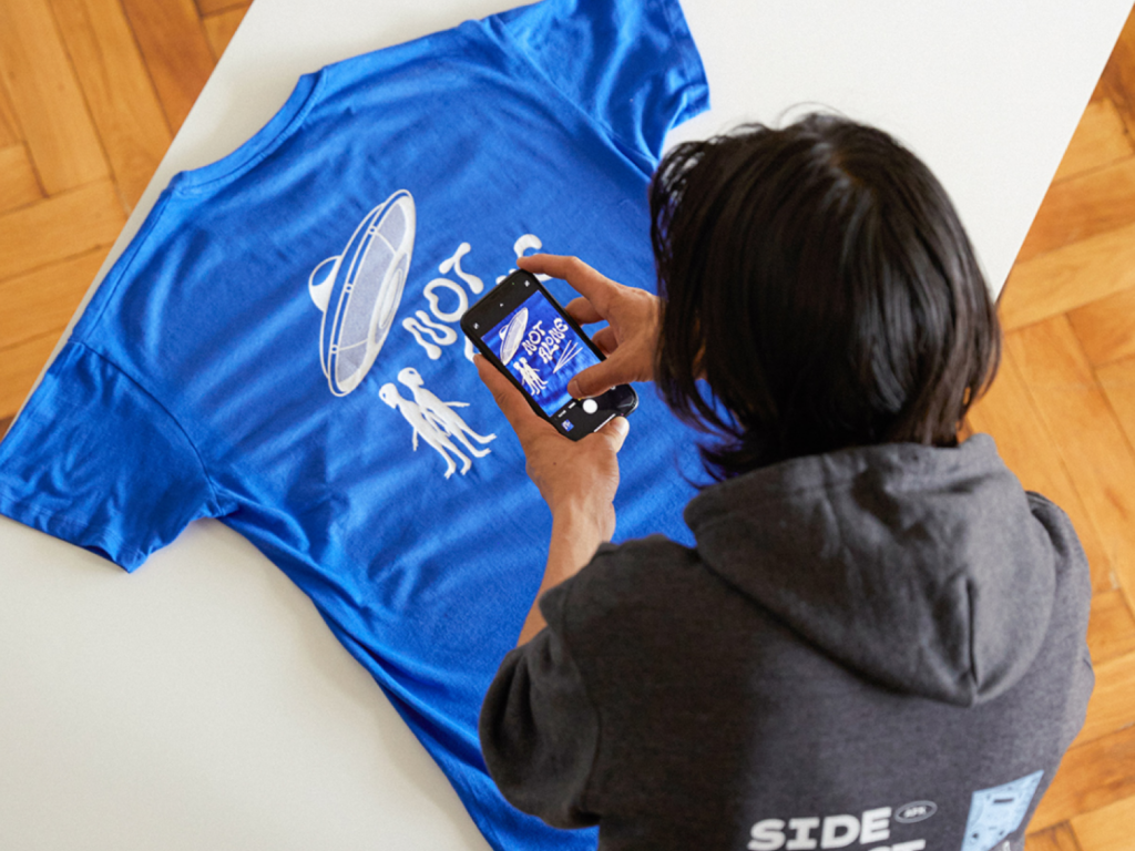 A man is taking a picture of a blue “Not alone” alien-themed t-shirt, using a smartphone.