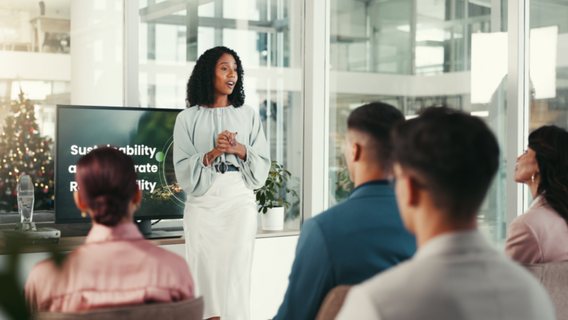 A woman stands confidently at the front, presenting to an engaged audience of diverse individuals seated in a conference room.
