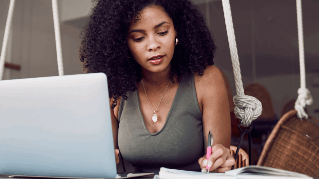 A woman is using a laptop and making notes in a notepad.