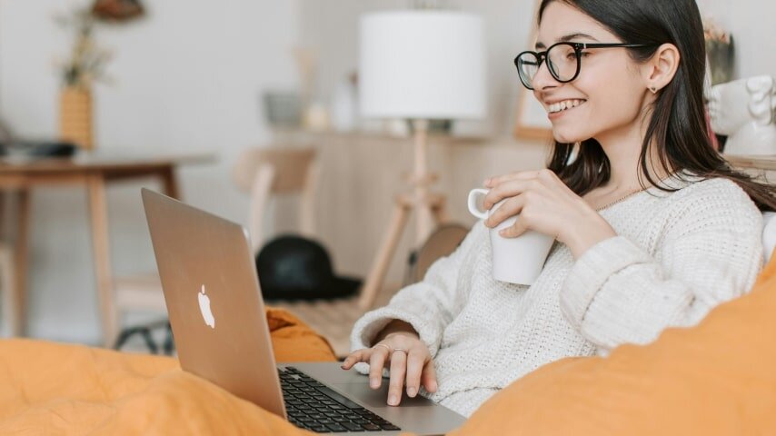 A woman drinking from a cup and looking at a laptop screen smiling.