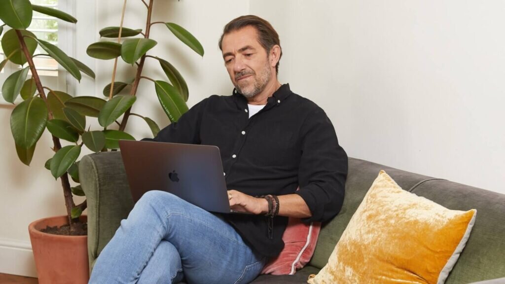 A man sitting on a couch with a laptop on his lap, working.