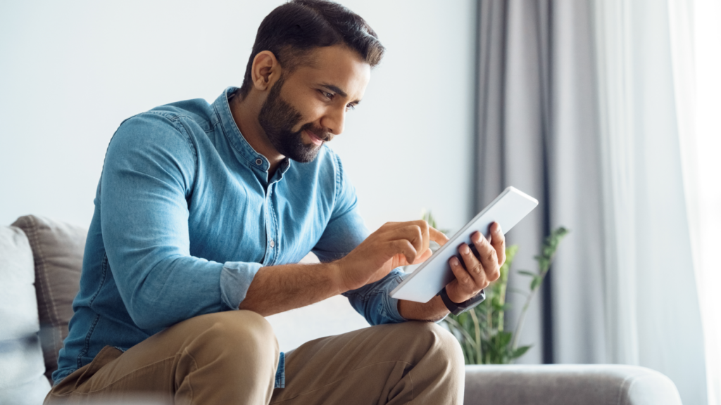 A man in a blue denim shirt sitting on a couch, looking at a tablet in his hands.