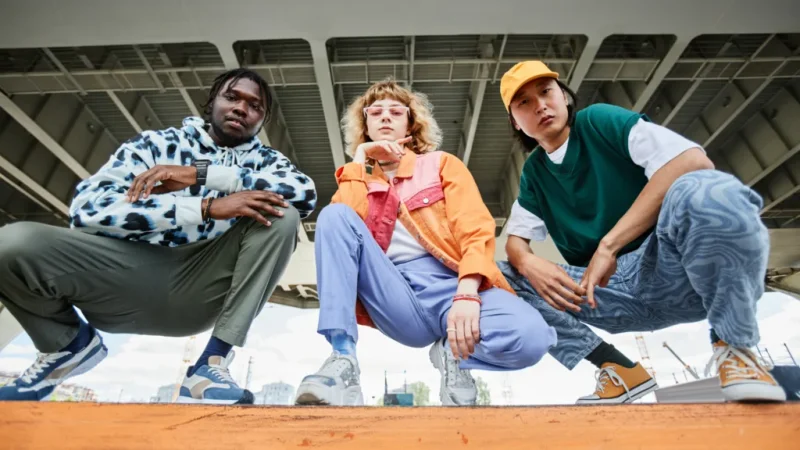 A group of people (man, woman, man), wearing fashionable clothes, squatting and looking at the camera.