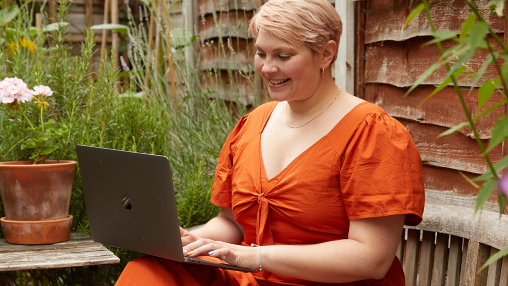A smiling young woman in an orange dress sitting outside and working using a laptop.